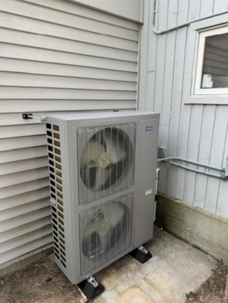 American Standard side-discharge heat pump outdoor unit on a concrete pad with vibration isolation, electrical disconnect and conduit run along the wall.